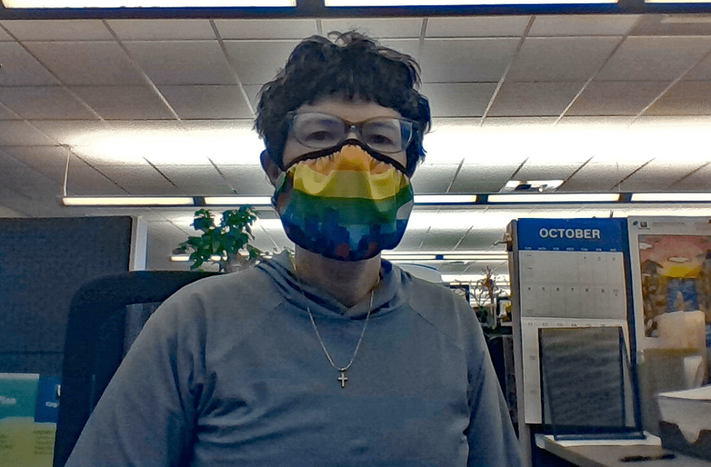 A woman wearing a rainbow colored mask sits at a desk in an office. The calendar behind her is set for October 2020, Disability Awareness Month. 